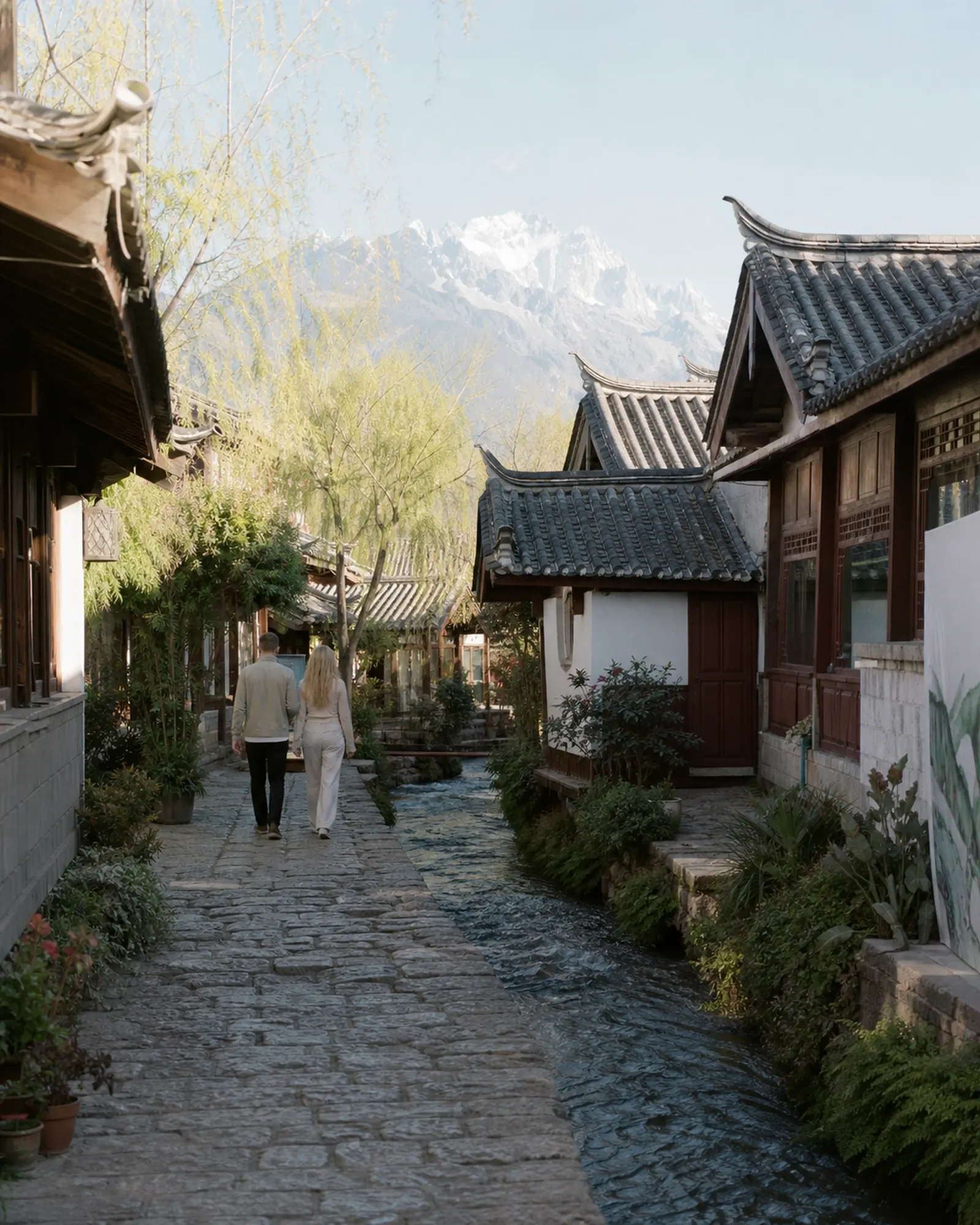 A couple walking a cobbled lane beside a stream in Lijiang old town, Jade Dragon Snow Mountain rising beyond the rooftops